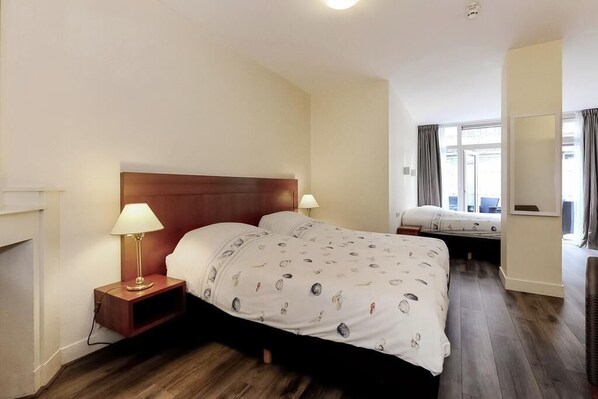 A hotel room with a wooden headboard bed, a bedside table with a lamp, a second bed in the background, and a large window with curtains. - Hotel Bor Scheveningen (The Hague)