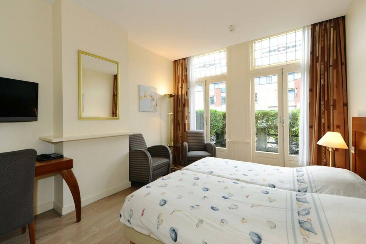 A hotel room with a bed, a desk, a chair, a TV, and large windows.