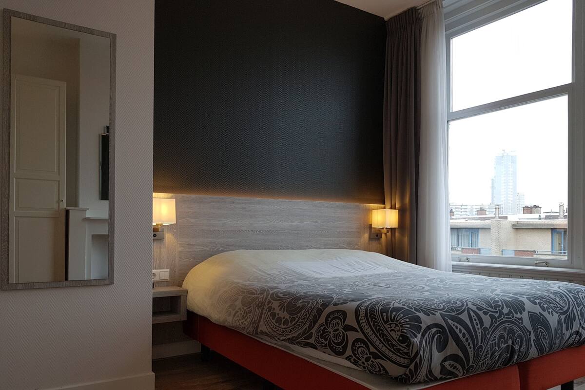 A hotel room with a large bed, a nightstand with a lamp, a mirror, and a window with a city view.