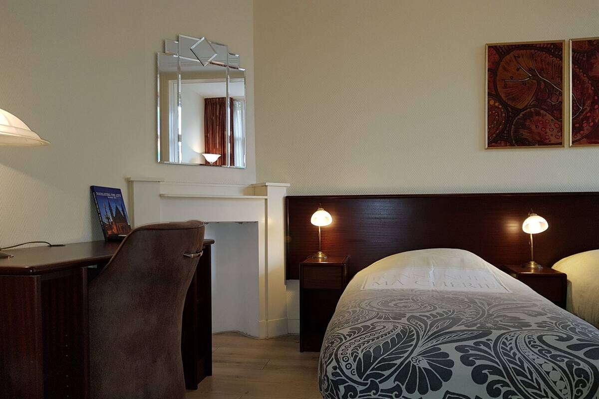A hotel room with a bed, two bedside tables with lamps, a desk with a chair, a mirror, and a framed picture on the wall.