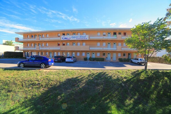 Ein Hotel mit zwei Etagen, an dem ein Schild mit der Aufschrift „Côte d'Azur Hotel“ zu sehen ist. Vor dem Hotel sind mehrere Autos geparkt, und auf der rechten Seite ist ein Baum sichtbar.