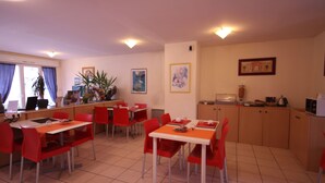 Un coin repas avec des chaises et des tables rouges, un espace cuisine avec des placards et une fenêtre avec des rideaux.