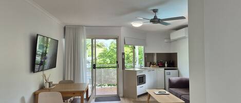 A modern kitchen with a built-in oven, a dining area with a wooden table and chairs, a sofa, and a ceiling fan.