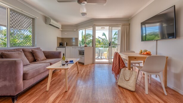 A modern living room with a sofa, coffee table, and TV. It features wooden flooring, a balcony with a view, and a kitchen area.