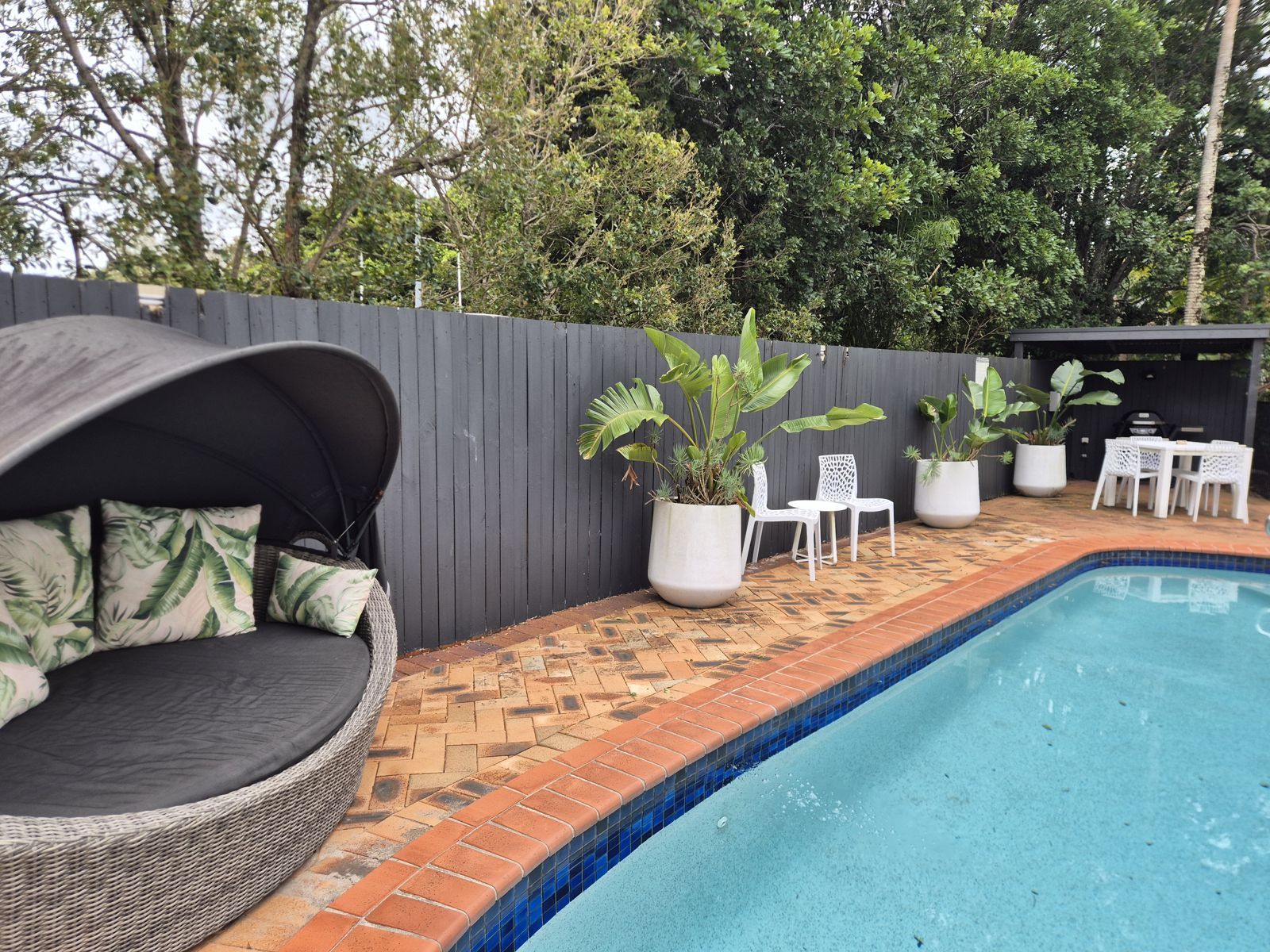 A swimming pool with a tiled edge, surrounded by a brick patio. There is a wicker lounge chair with cushions, white chairs, and potted plants.