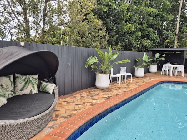 A swimming pool with a tiled edge, surrounded by a brick patio. There is a wicker lounge chair with cushions, white chairs, and potted plants.