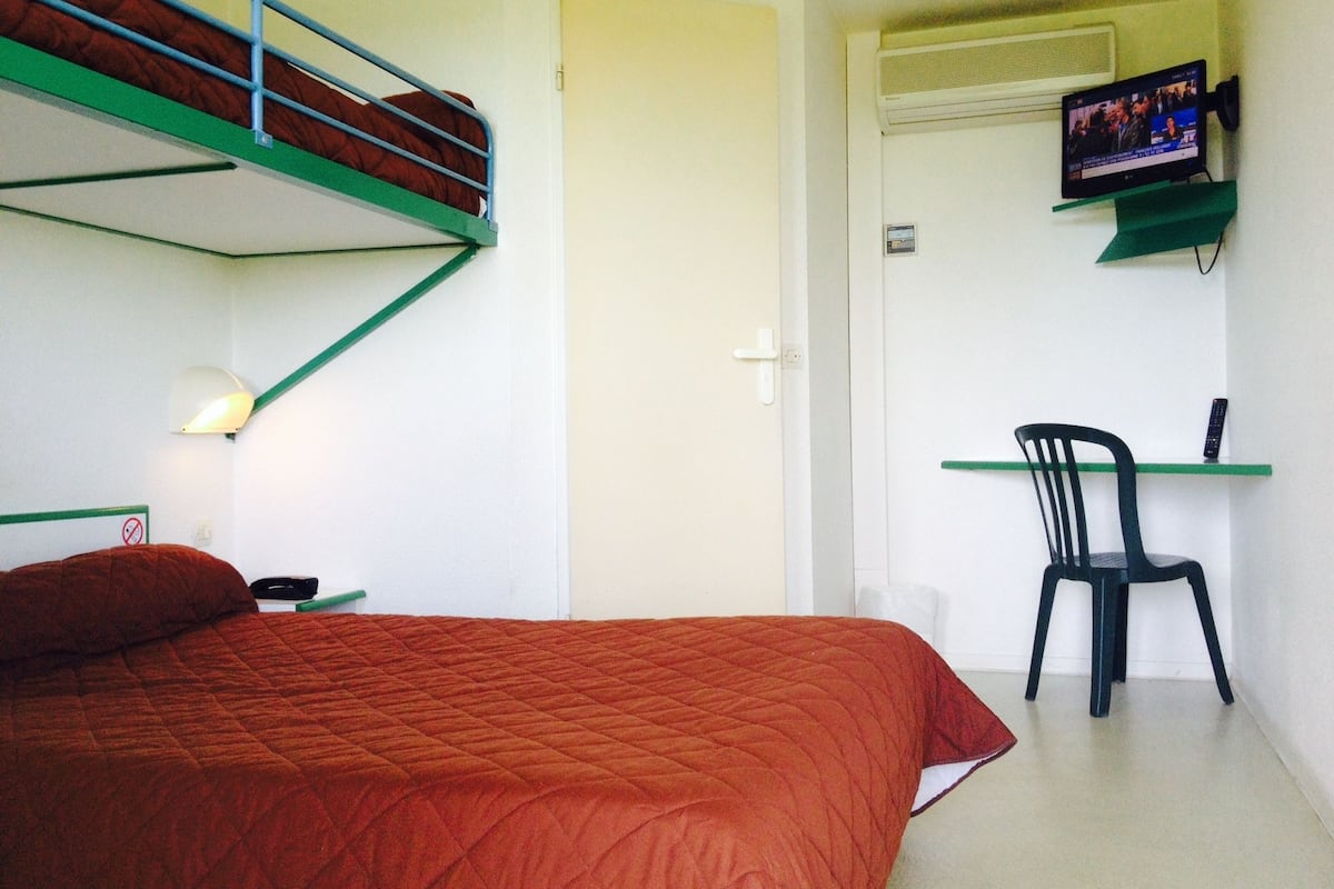 A dormitory room with a bunk bed, a desk with a chair, a television, and a bed with a red blanket.