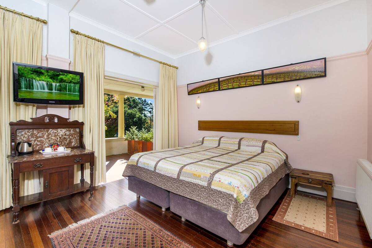 A bedroom with a large bed, a wooden nightstand, a television on a dresser, and a window with curtains.