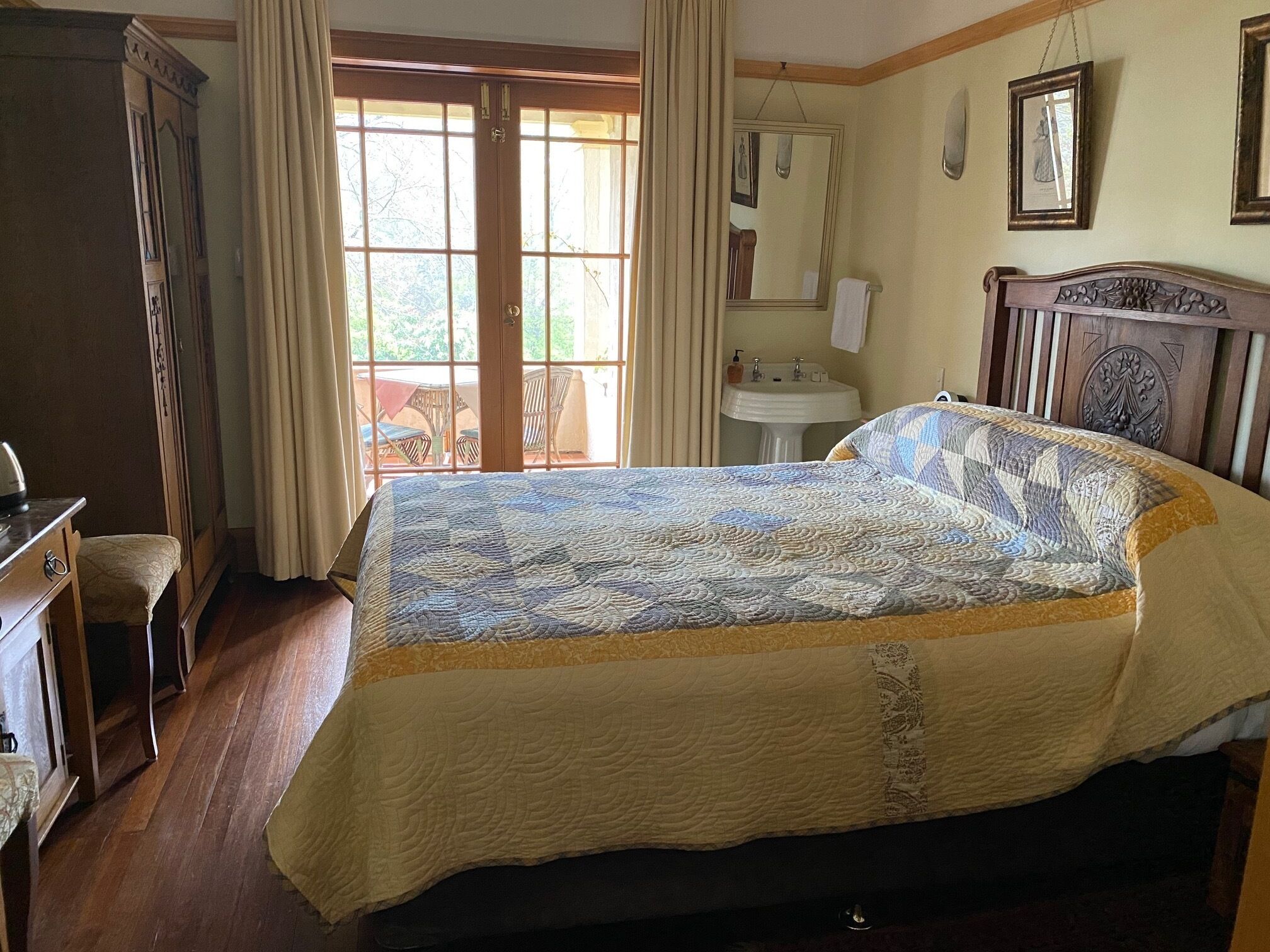 Un dormitorio con una cama grande, un aparador de madera, un escritorio, una silla, un ventanal con cortinas, un lavamanos y un espejo.