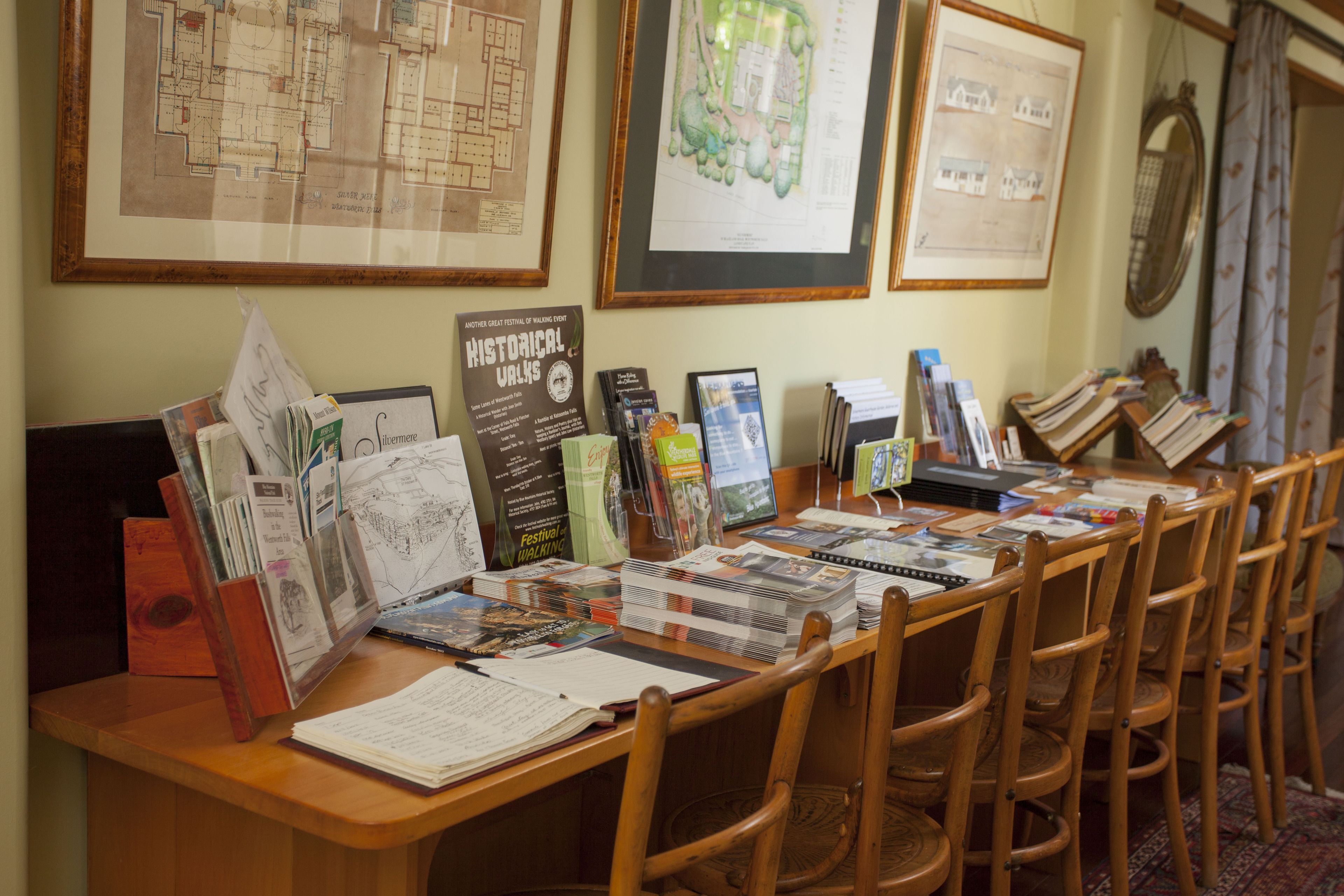 Un mostrador de recepción con diversos folletos informativos, planos arquitectónicos enmarcados y un cartel anunciando el festival de Paseos Históricos.