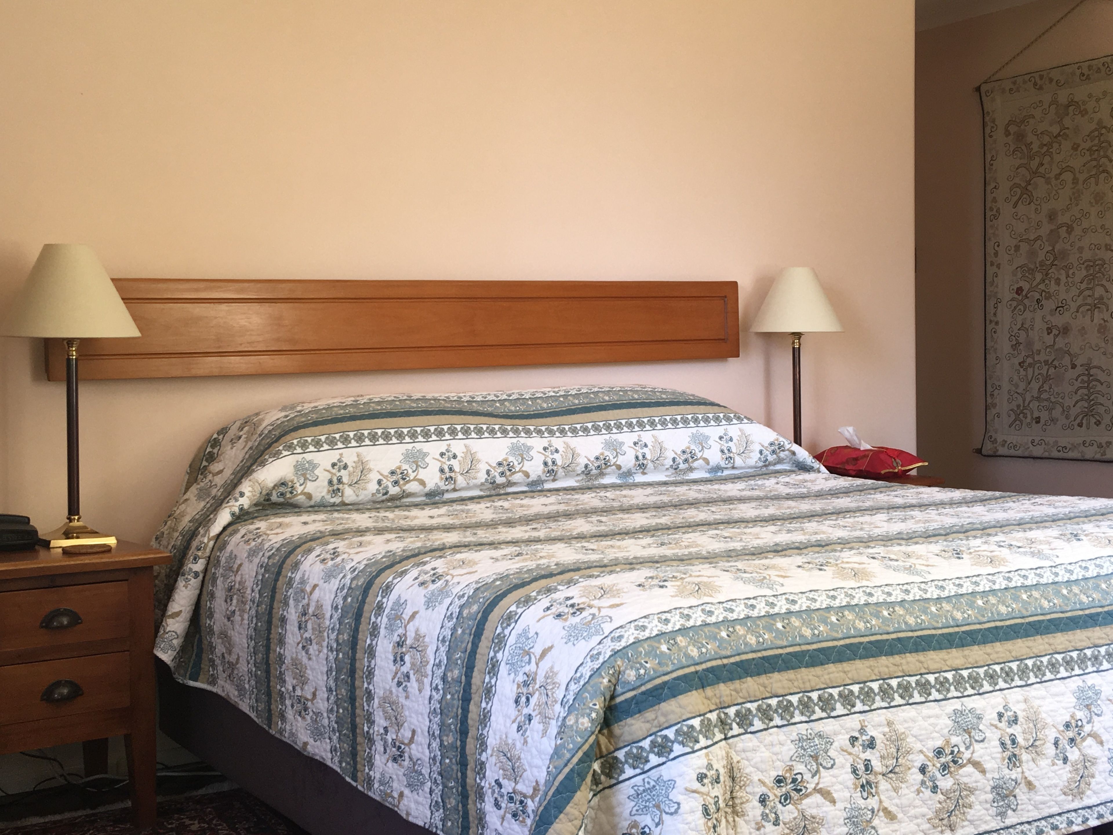 A bedroom with a wooden headboard, a floral-patterned bedspread, two bedside lamps, a bedside table with a lamp, and a red object on the floor.