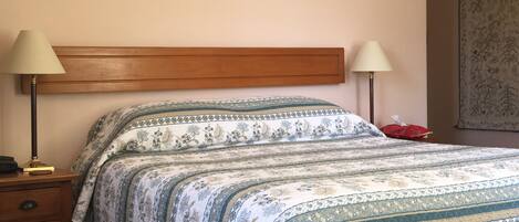 A bedroom with a wooden headboard, a floral-patterned bedspread, two bedside lamps, a bedside table with a lamp, and a red object on the floor.
