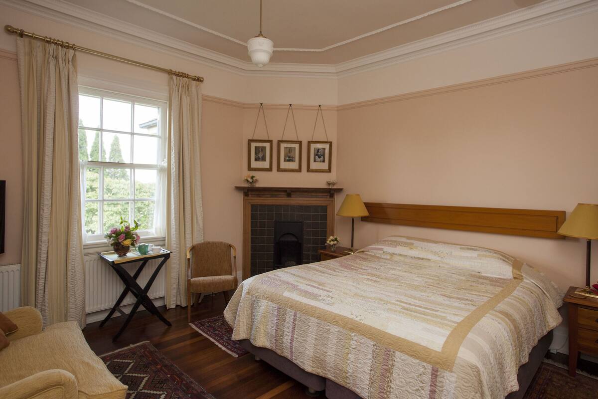 A bedroom with a large bed, a fireplace, a window with curtains, a chair, a small table with a vase, and a lamp.