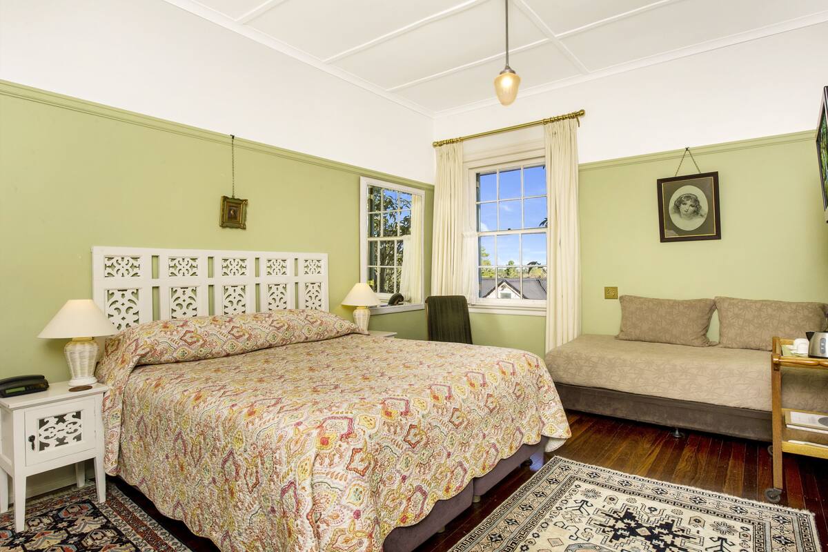 A bedroom with a large bed, a smaller sofa, a nightstand with a lamp, a chair, and a window with curtains.
