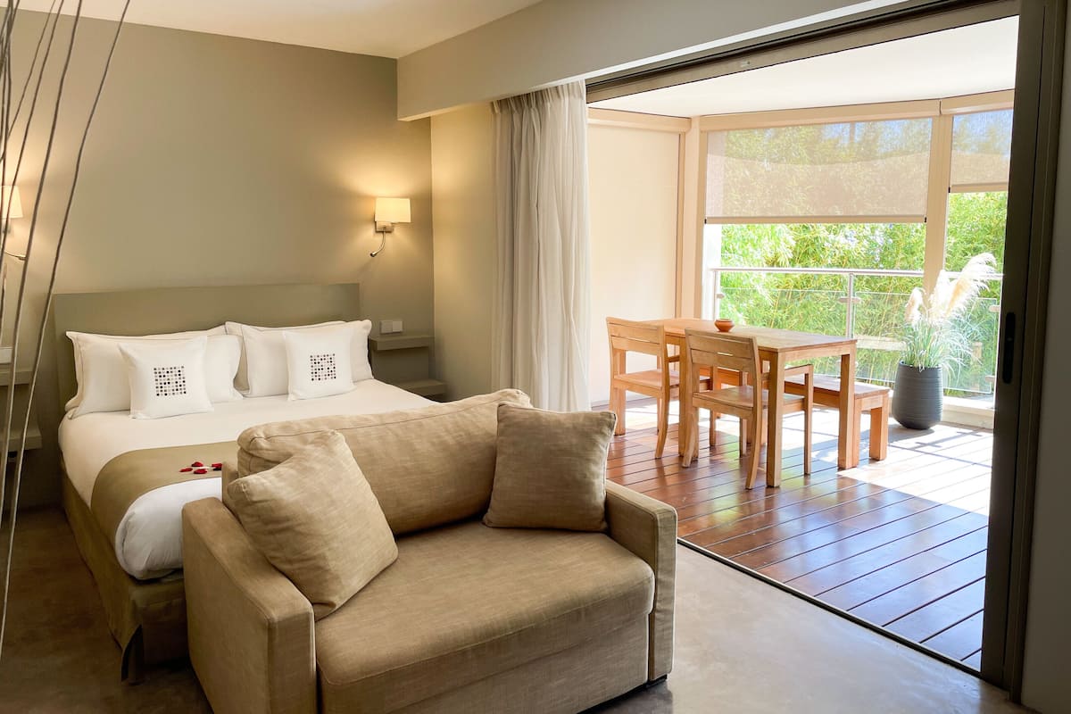 A hotel room with a bed, a sofa, a table with chairs, and a balcony with a view of greenery.