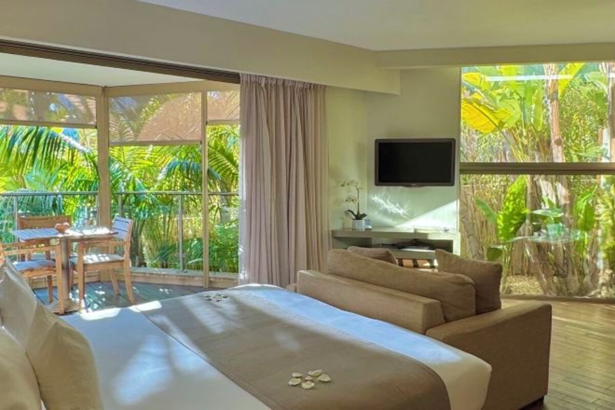 Quarto de hotel com cama, televisão, varanda com mesa e cadeiras, e vista para a vegetação tropical.