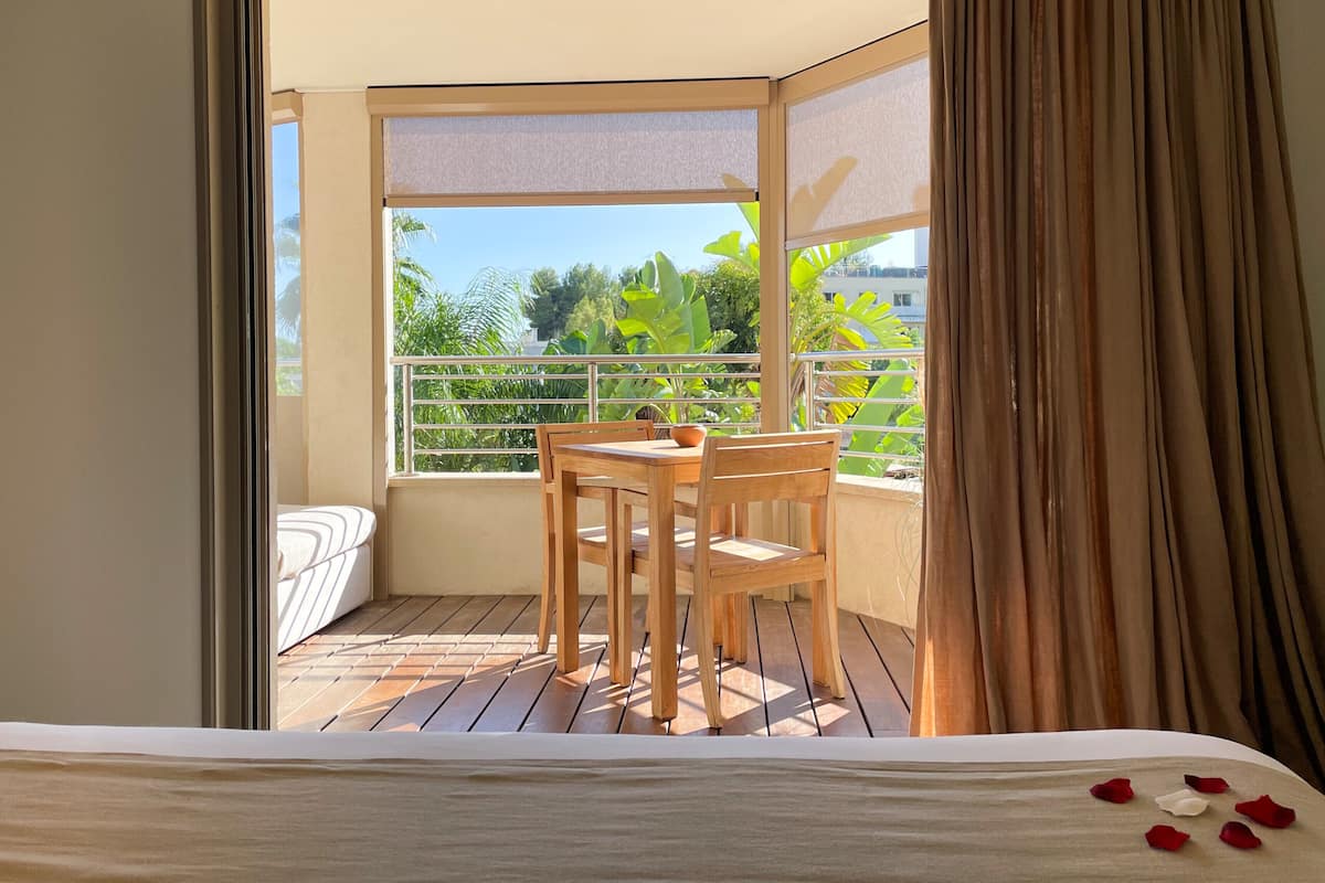 A balcony with a wooden table and chairs, a view of greenery, and a white bed with a white sheet.