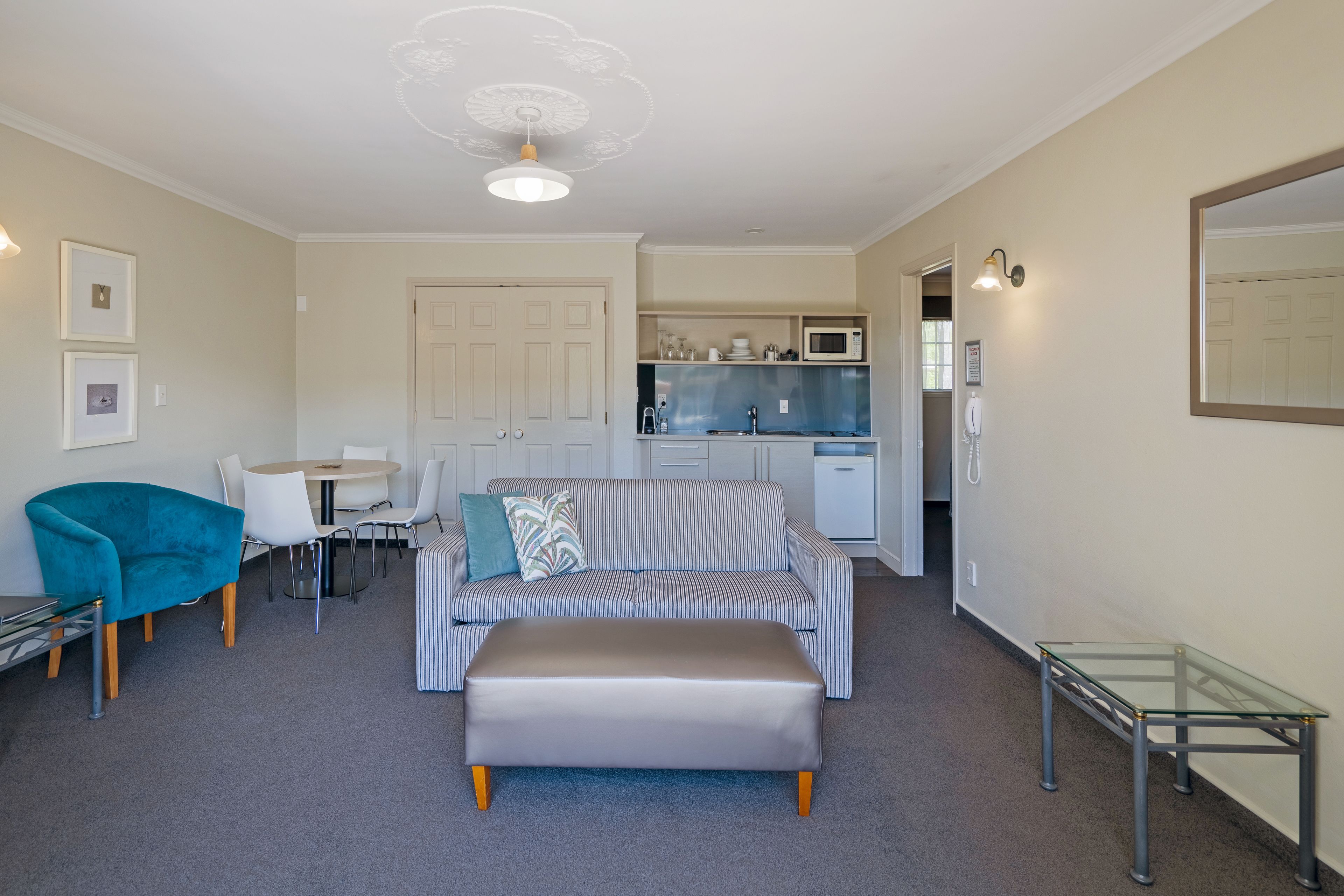 A living room with a striped sofa, a teal armchair, a glass coffee table, and a kitchenette in the background.