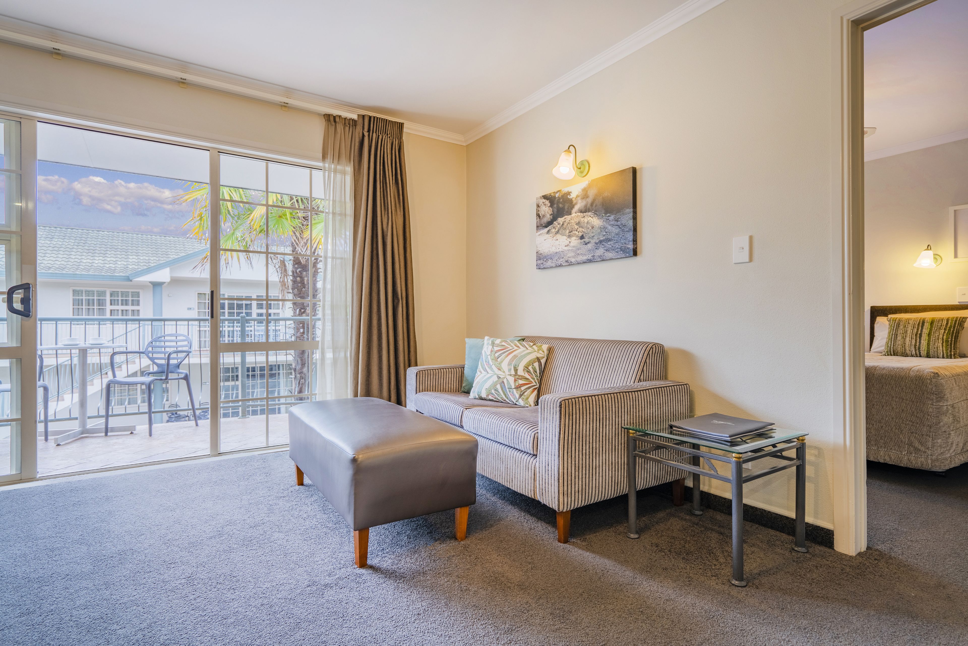 A living room with a sofa, ottoman, and a table with a laptop. There is a sliding glass door leading to a balcony with a chair.