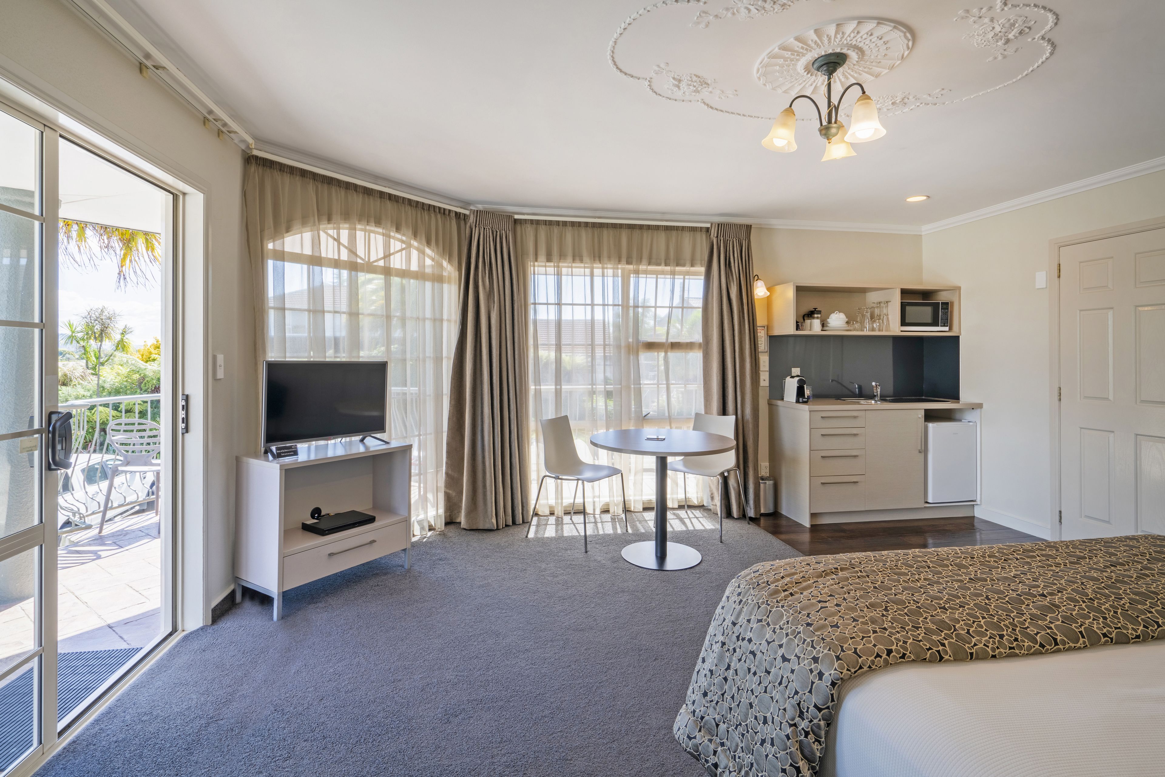 Ein Hotelzimmer mit Bett, Fernseher, Schreibtisch mit Stuhl, einer kleinen Küchenzeile und einem Balkon mit Tisch und Stühlen.