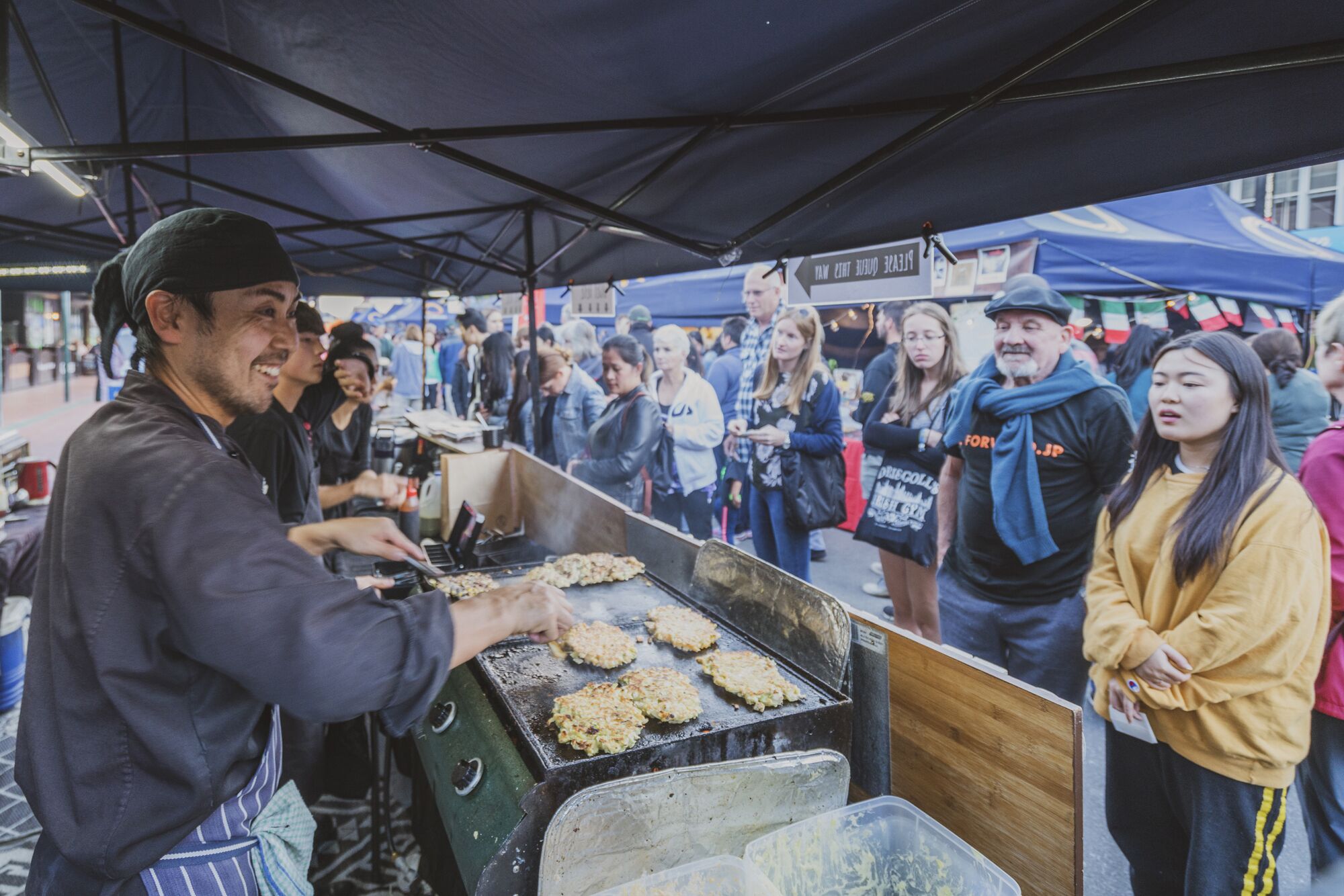 Un vendedor ambulante de comida asando alimentos debajo de una carpa, mientras una multitud observa.