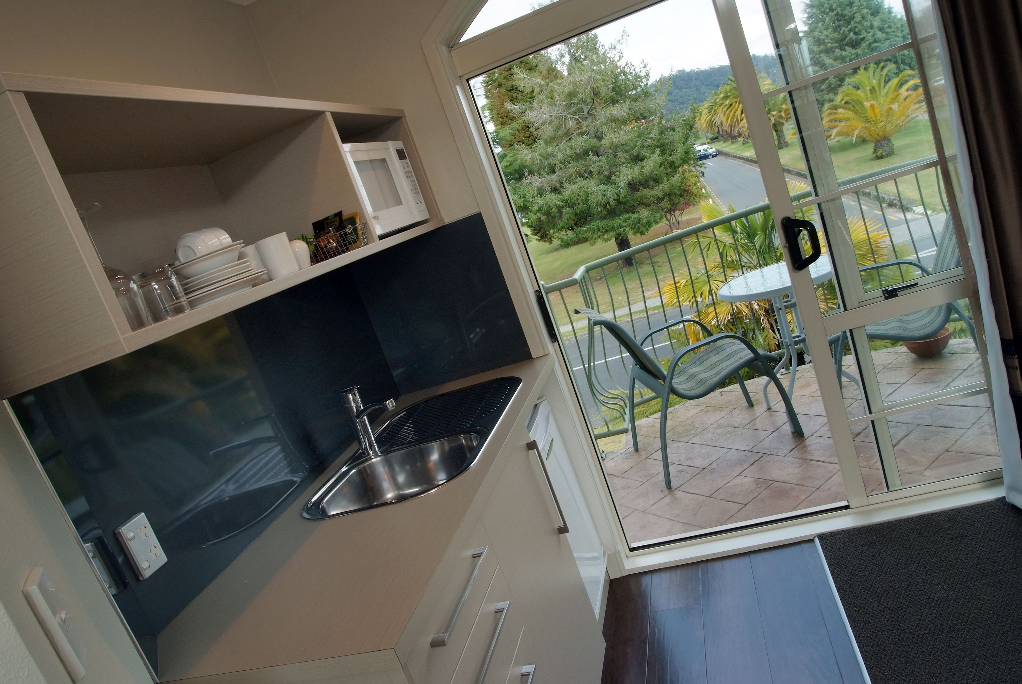A compact kitchen with a sink and a microwave, a balcony with a table and chairs, and a view of a landscaped garden.
