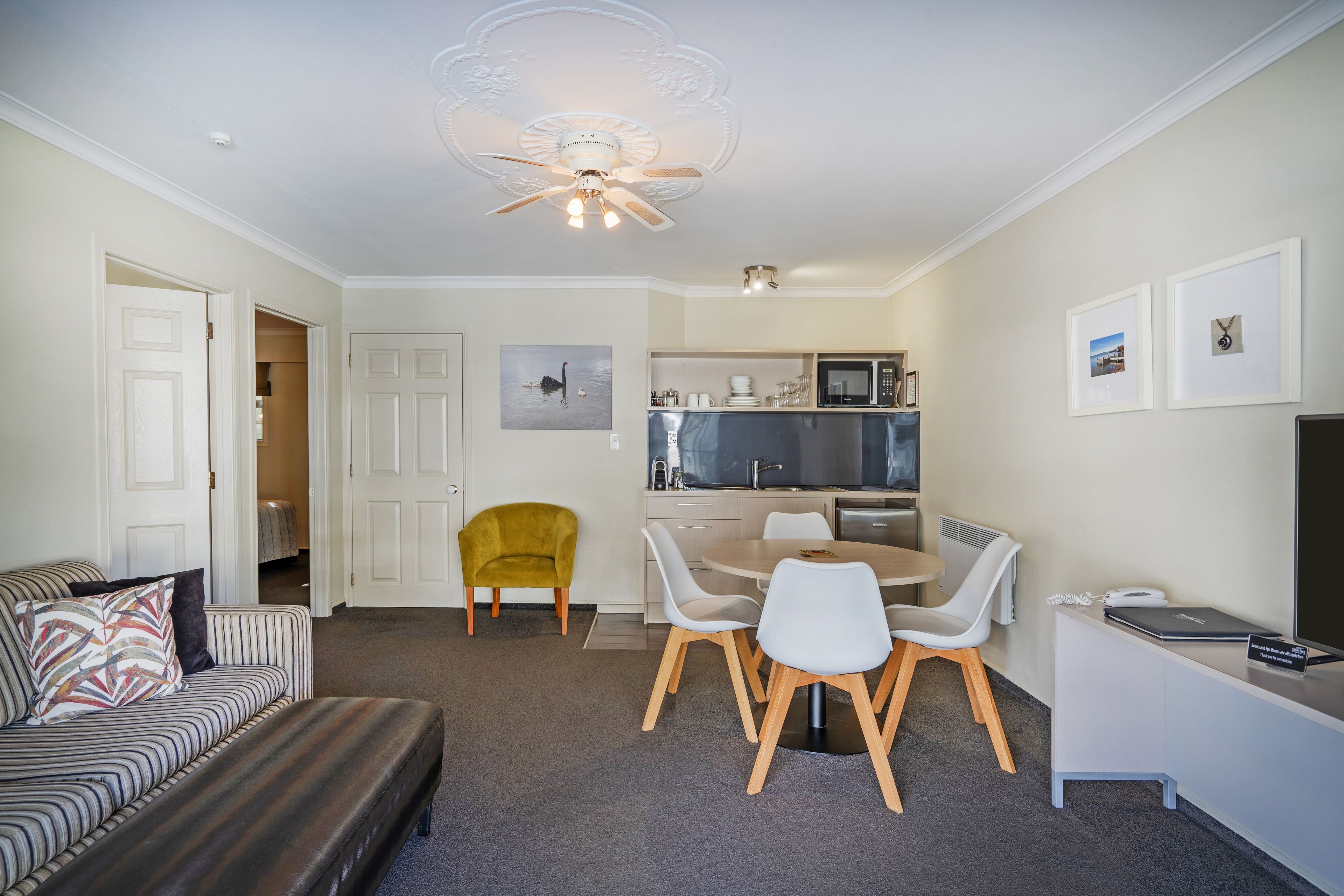 A modern living room with a sofa, a dining table with chairs, a kitchen area with a microwave, and a ceiling fan.