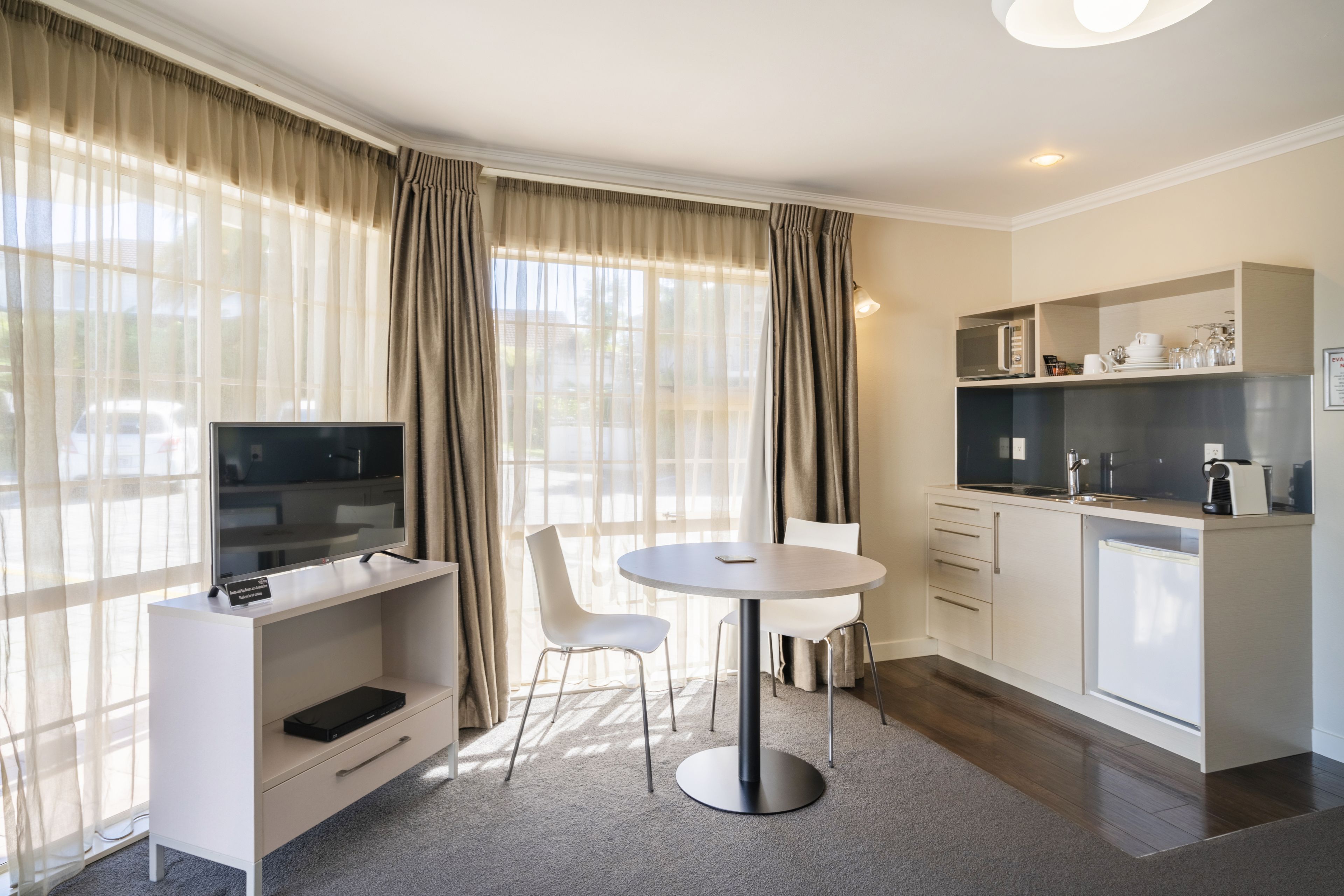 Una habitación de hotel moderna con televisión de pantalla plana, una mesa de comedor con sillas, kitchenette y ventanas amplias con cortinas.
