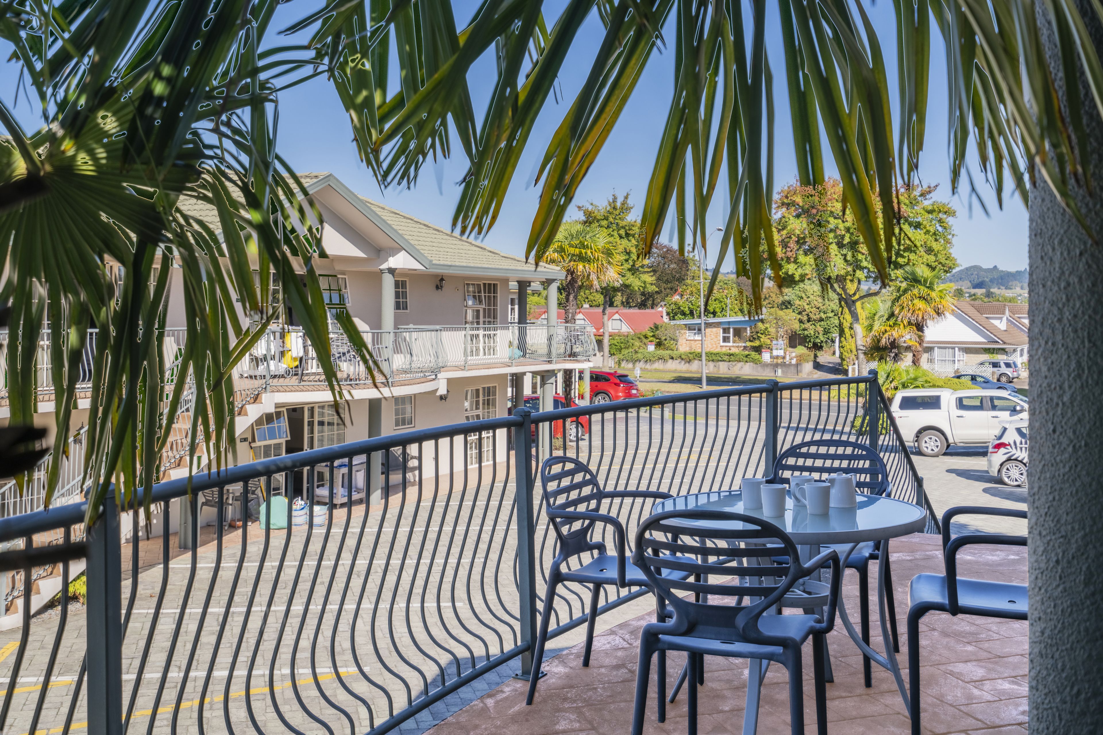 A balcony with a metal railing, outdoor seating with chairs and a table, and a view of a residential area with houses and parked cars.