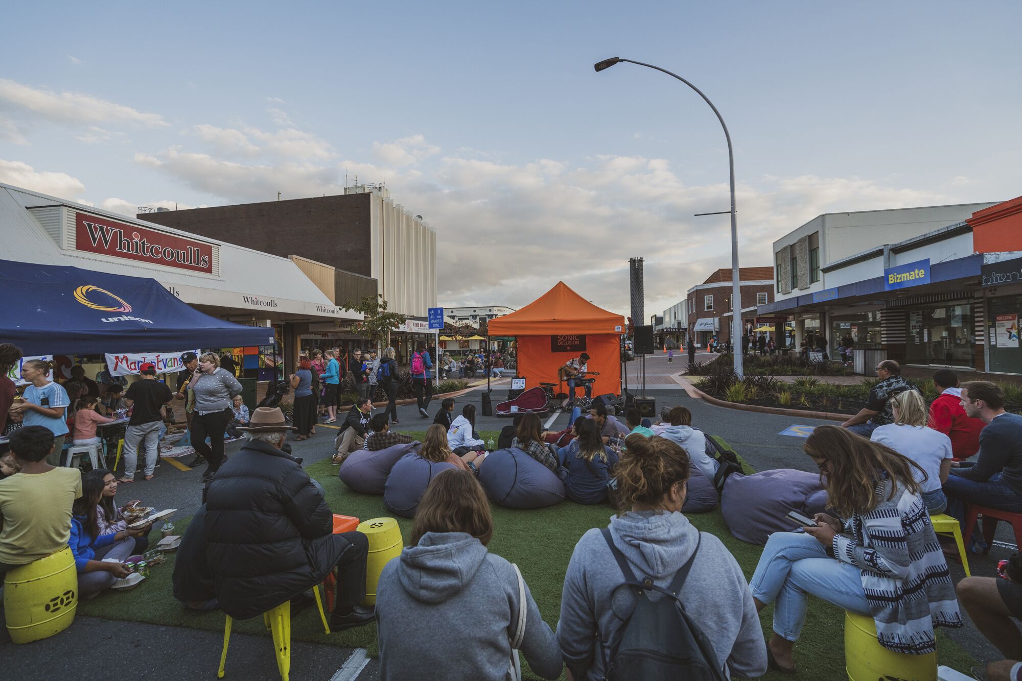 A group of people sitting on bean bags and chairs in an outdoor setting with a stage in the center.