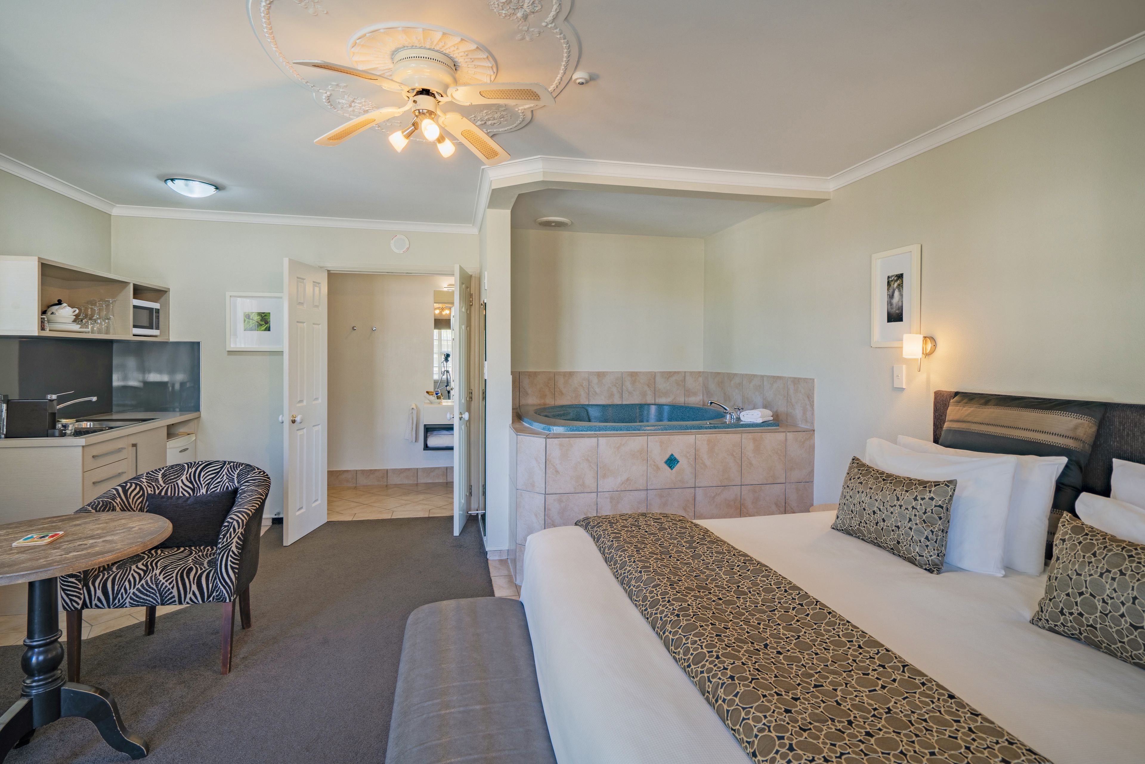 A hotel room with a bed, a desk, a chair, a ceiling fan, a bathroom with a bathtub, and a kitchenette.