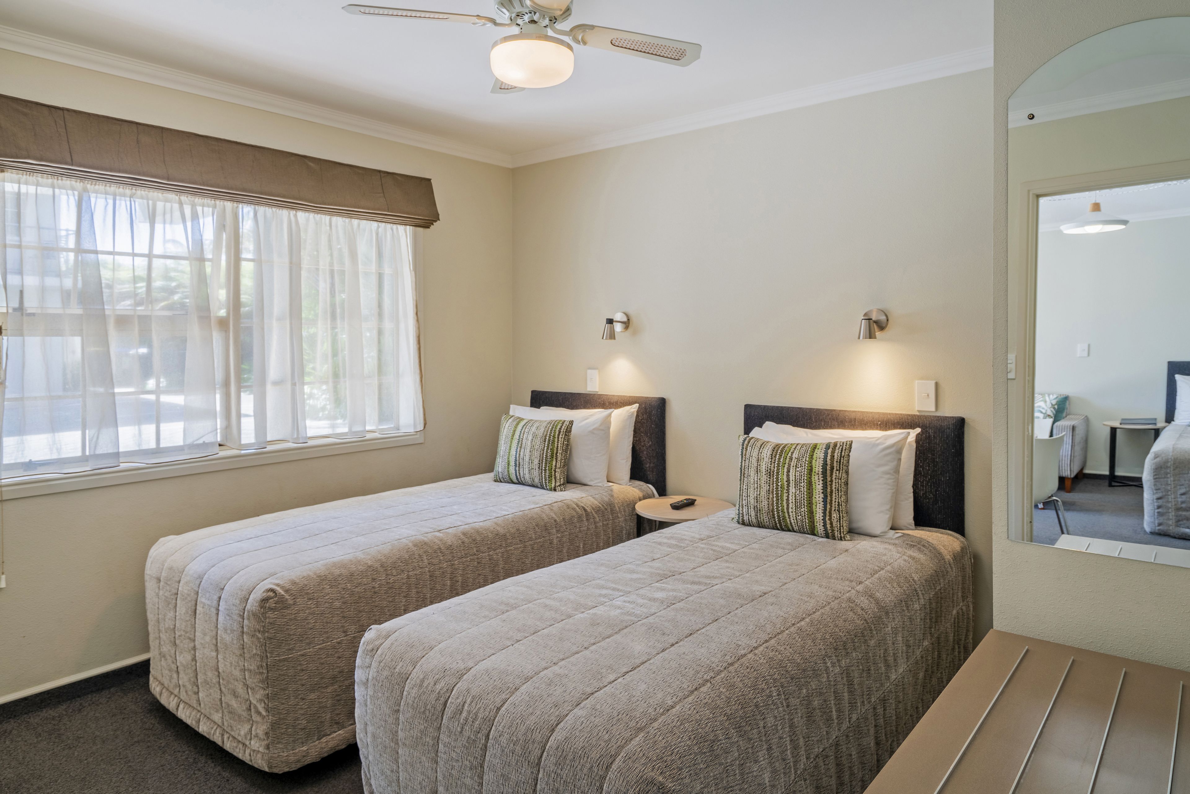 A hotel room with two beds, a ceiling fan, a window with curtains, and a mirror reflecting the room.