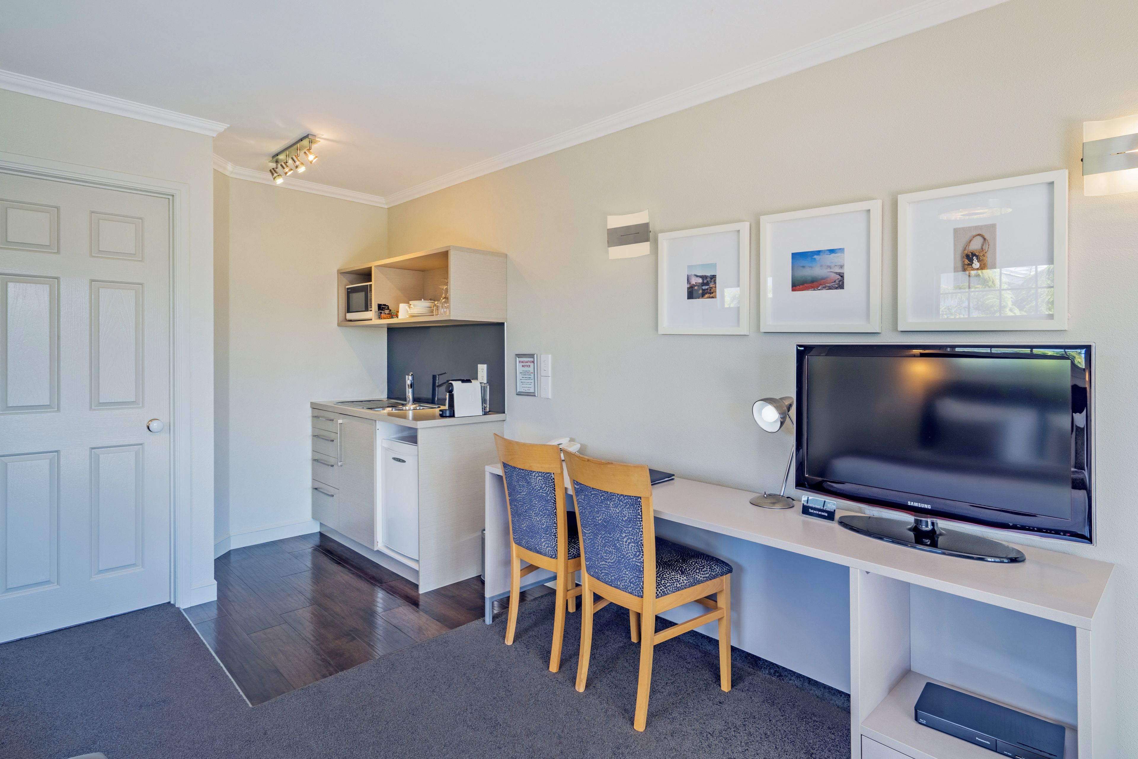 A compact kitchen with a microwave, sink, and cabinets, a dining area with two chairs, a flat-screen TV, and framed pictures on the wall.