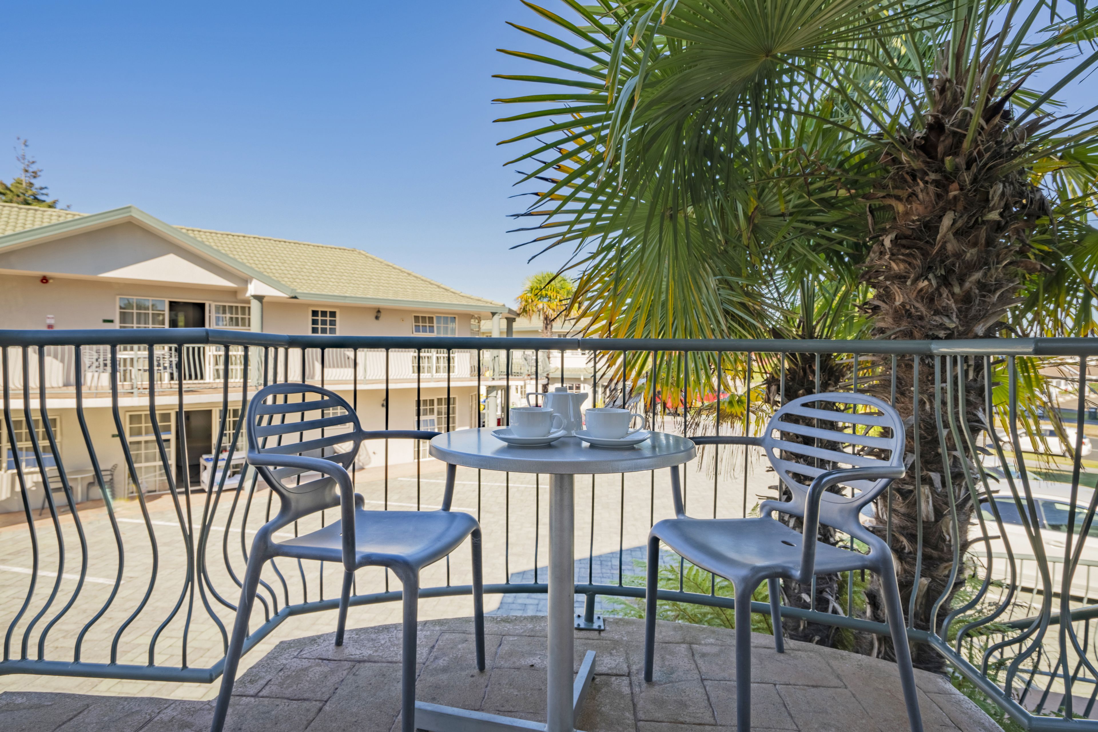A balcony with two chairs and a table, a palm tree, and a building in the background.