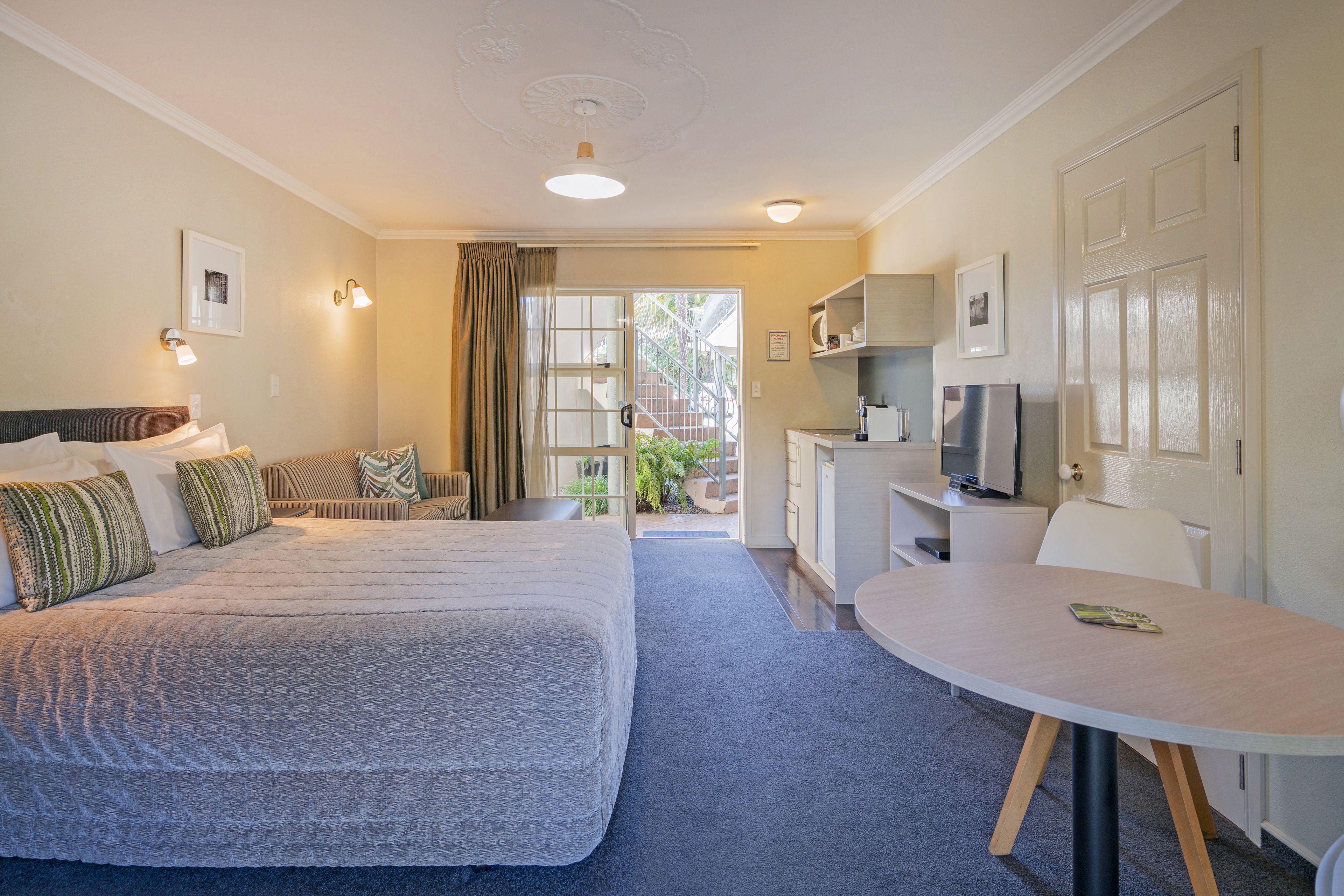 A hotel room with a large bed, a desk, a chair, and a small table. There is a door leading to a balcony.
