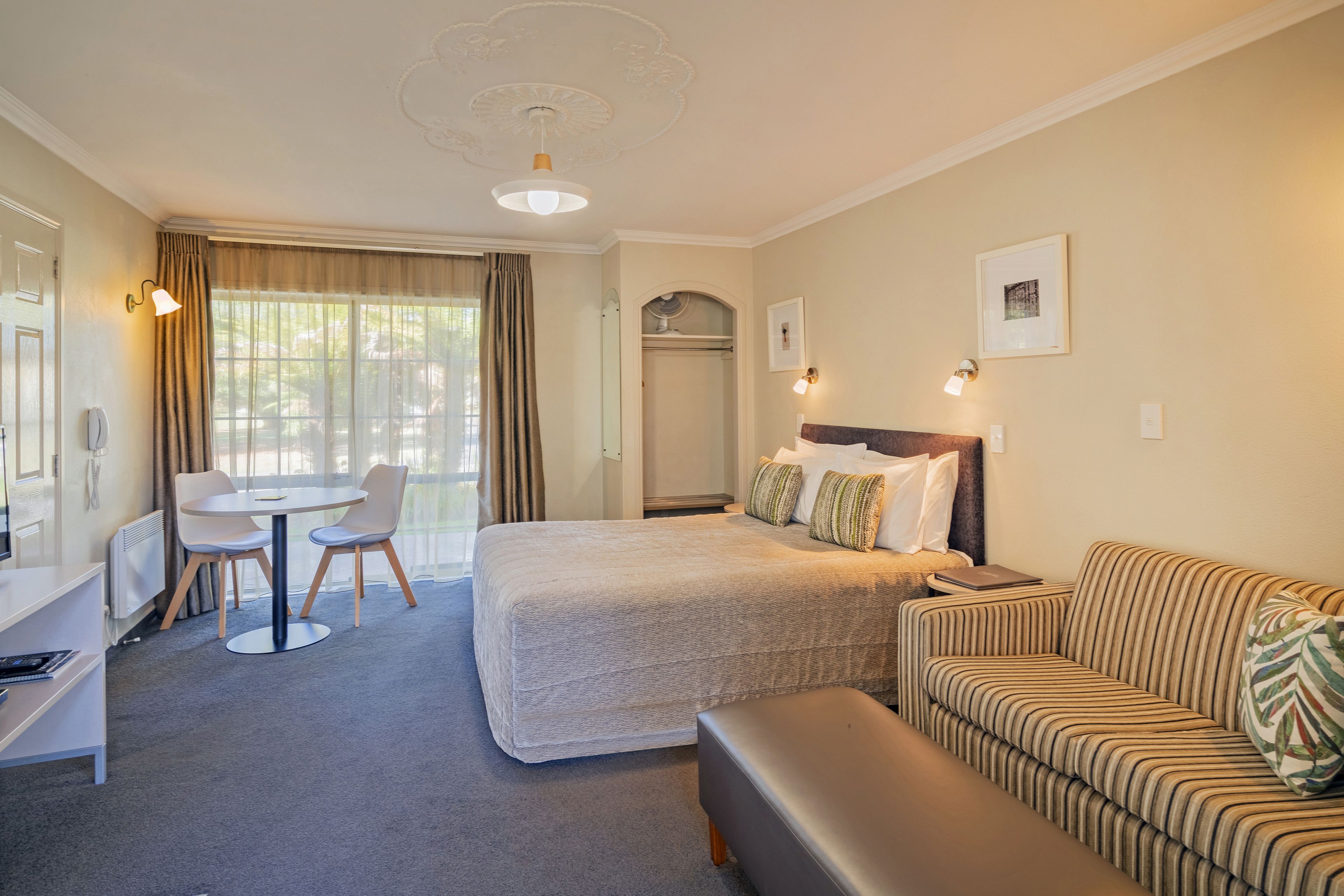 A hotel room with a bed, a sofa, a desk, and a chair. There is a window with curtains, a door, and a coat rack.