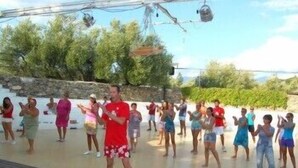 Un grupo de personas bailando una rutina al aire libre sobre una terraza de madera, con un muro de piedra y vegetación de fondo.