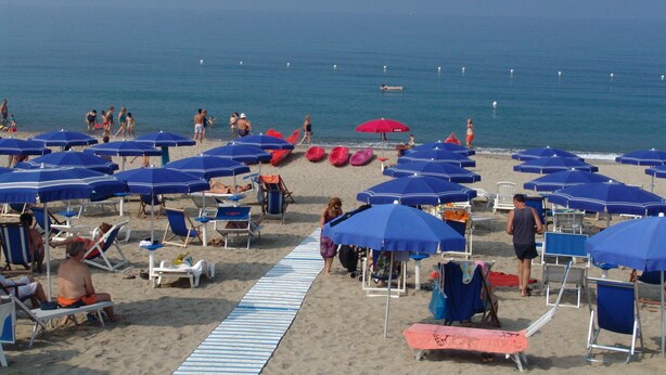Una playa con hileras de sombrillas azules y rojas, sillas de playa y un paseo marítimo arenoso.