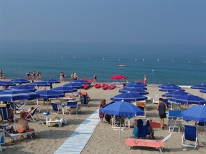 Una playa con hileras de sombrillas azules y rojas, sillas de playa y un paseo marítimo arenoso.