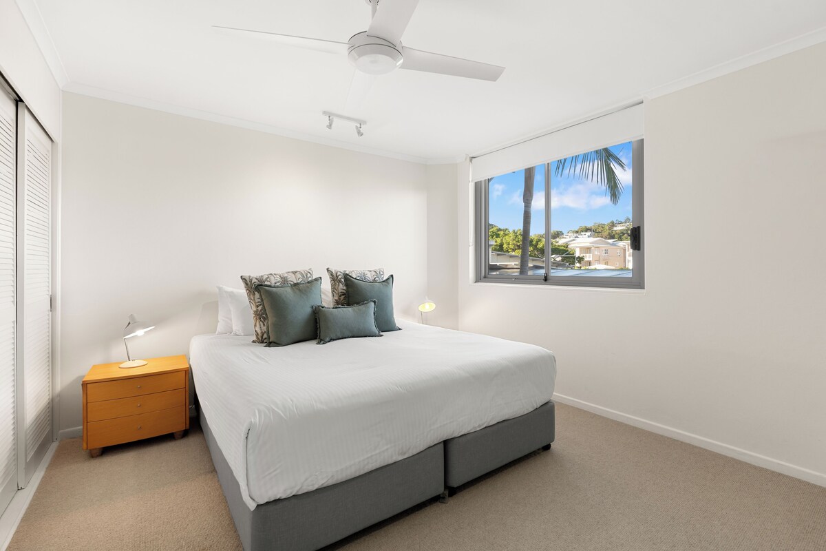 A bedroom with a bed, nightstand, ceiling fan, window, and view of a residential area.