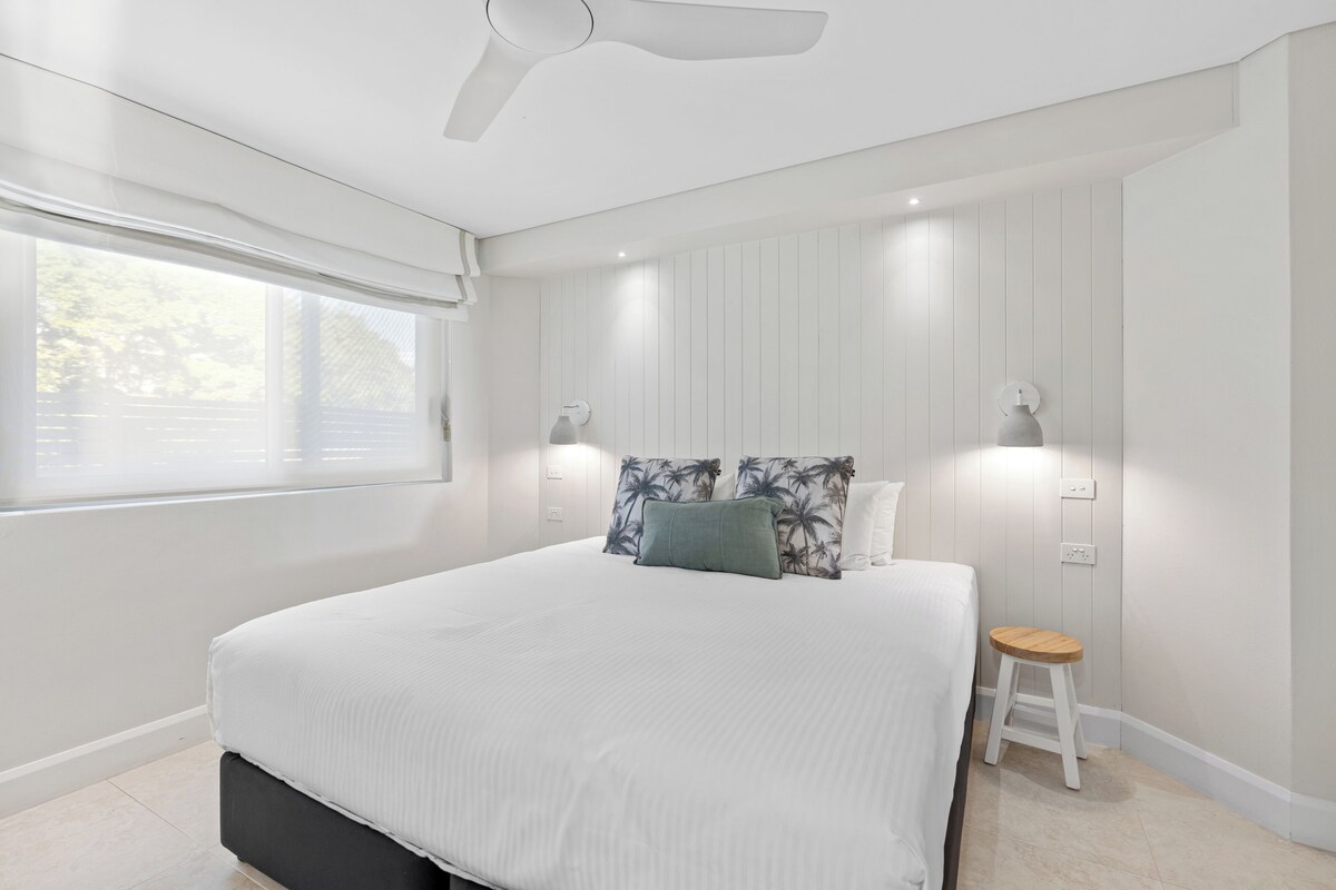 A bedroom with a large bed, a ceiling fan, two wall-mounted lights, a window with blinds, and a small stool.