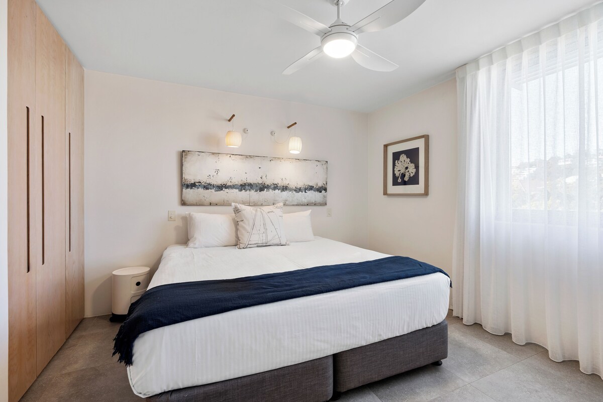 A modern bedroom with a large bed, a ceiling fan, and a view of the outdoors through sheer curtains.