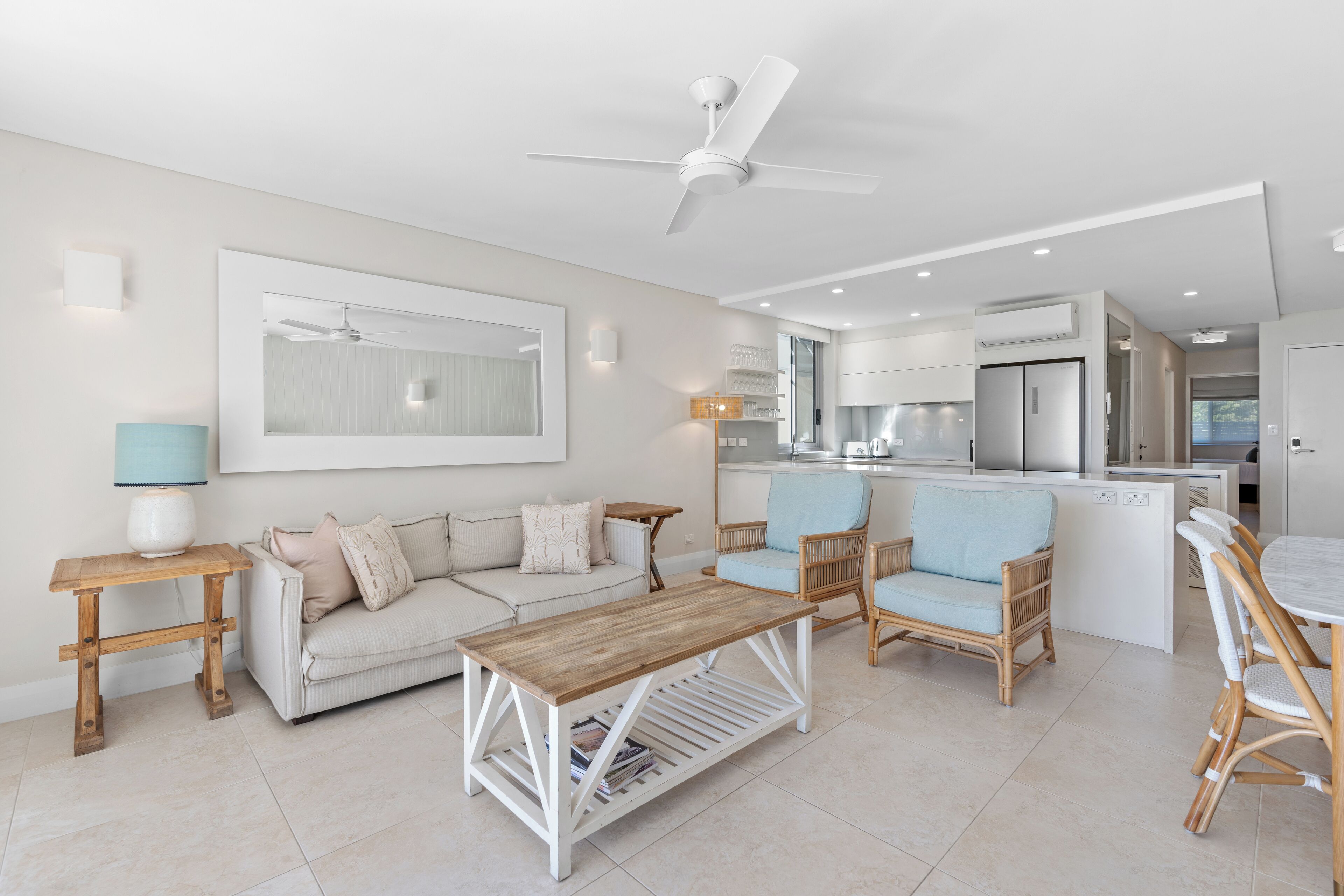 A modern living room with a sofa, coffee table, and wicker chairs. There is a kitchen in the background with a refrigerator and a dining area with a table and chairs.