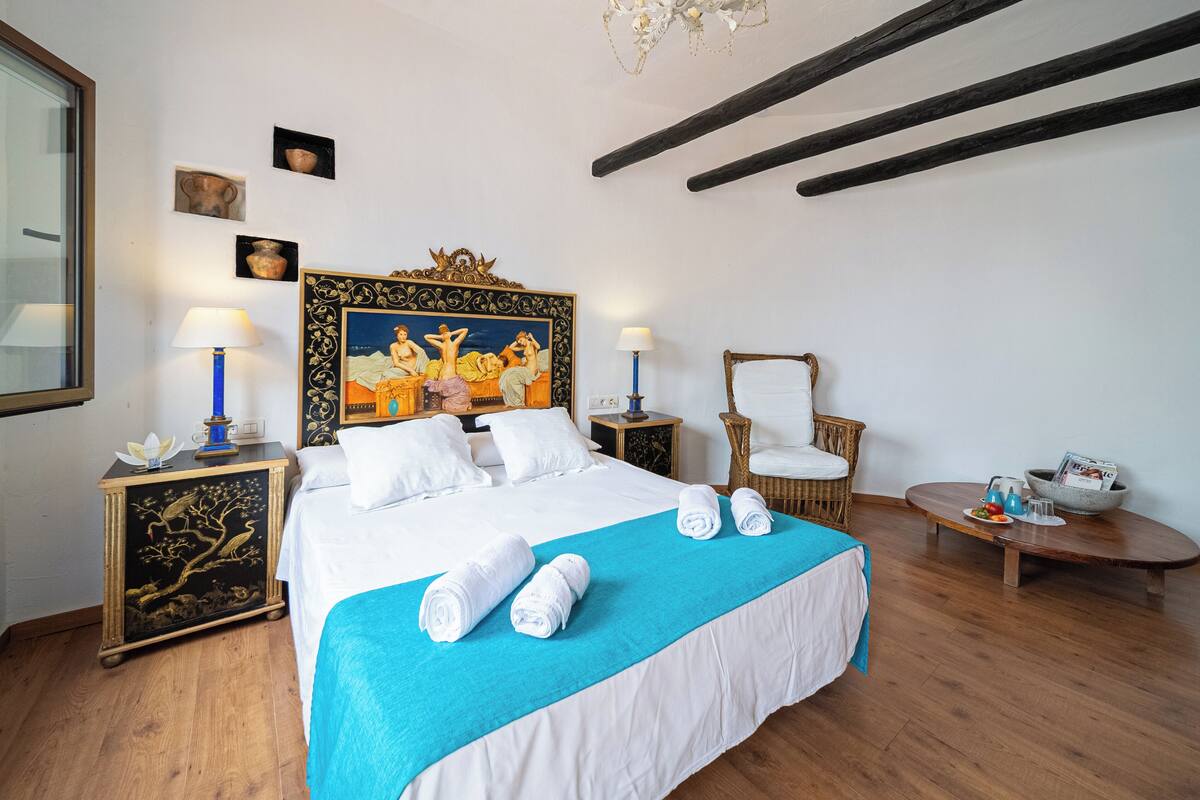 Una cama bien hecha con sábanas blancas y una manta azul, una mesita de noche de madera con una lámpara, una silla de mimbre y una pequeña mesa con un cuenco y una taza.