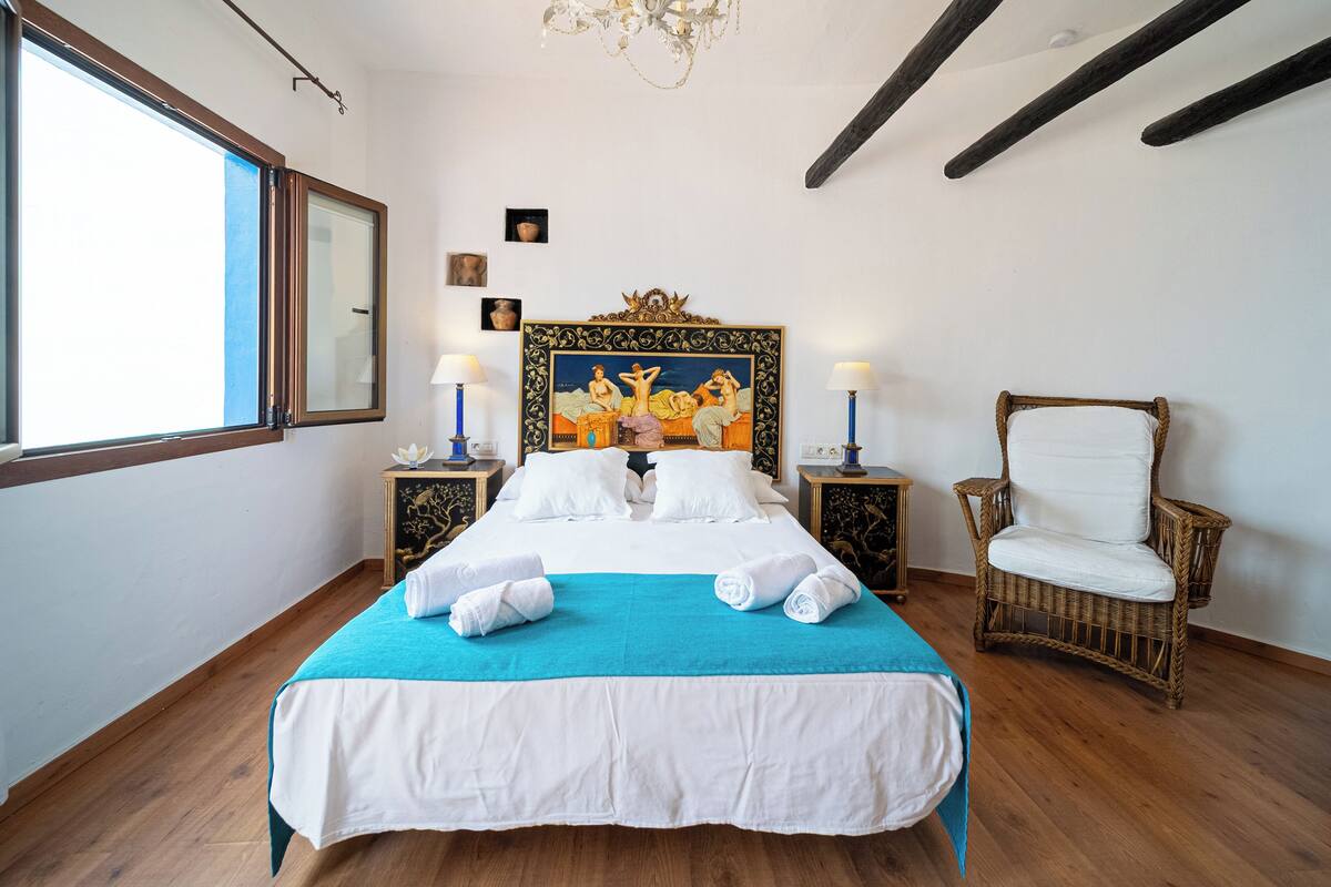 Una cama bien tendida con sábanas blancas y un detalle azul, una silla de mimbre y un ventanal con vistas.