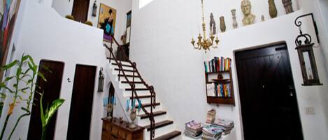 Una escalera conduce a un segundo piso, rodeada de paredes blancas, un candelabro y diversos objetos decorativos.