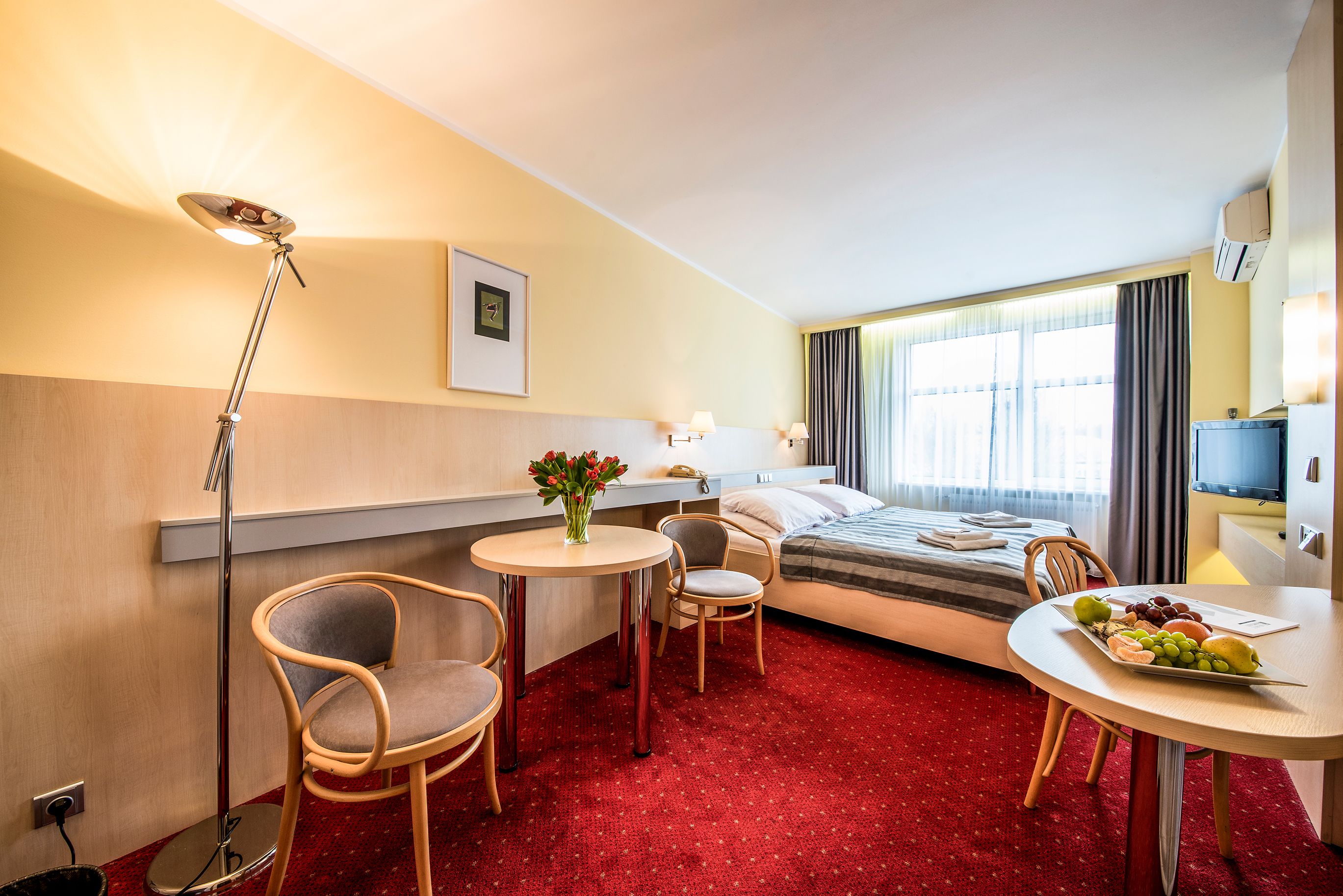 A hotel room with a bed, a desk, two chairs, a small table with fruit, a lamp, and a TV.