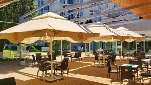An outdoor patio with multiple tables and chairs under large umbrellas, a modern building in the background, and a grassy area with a playground. - Hotel Jana (Prerov)