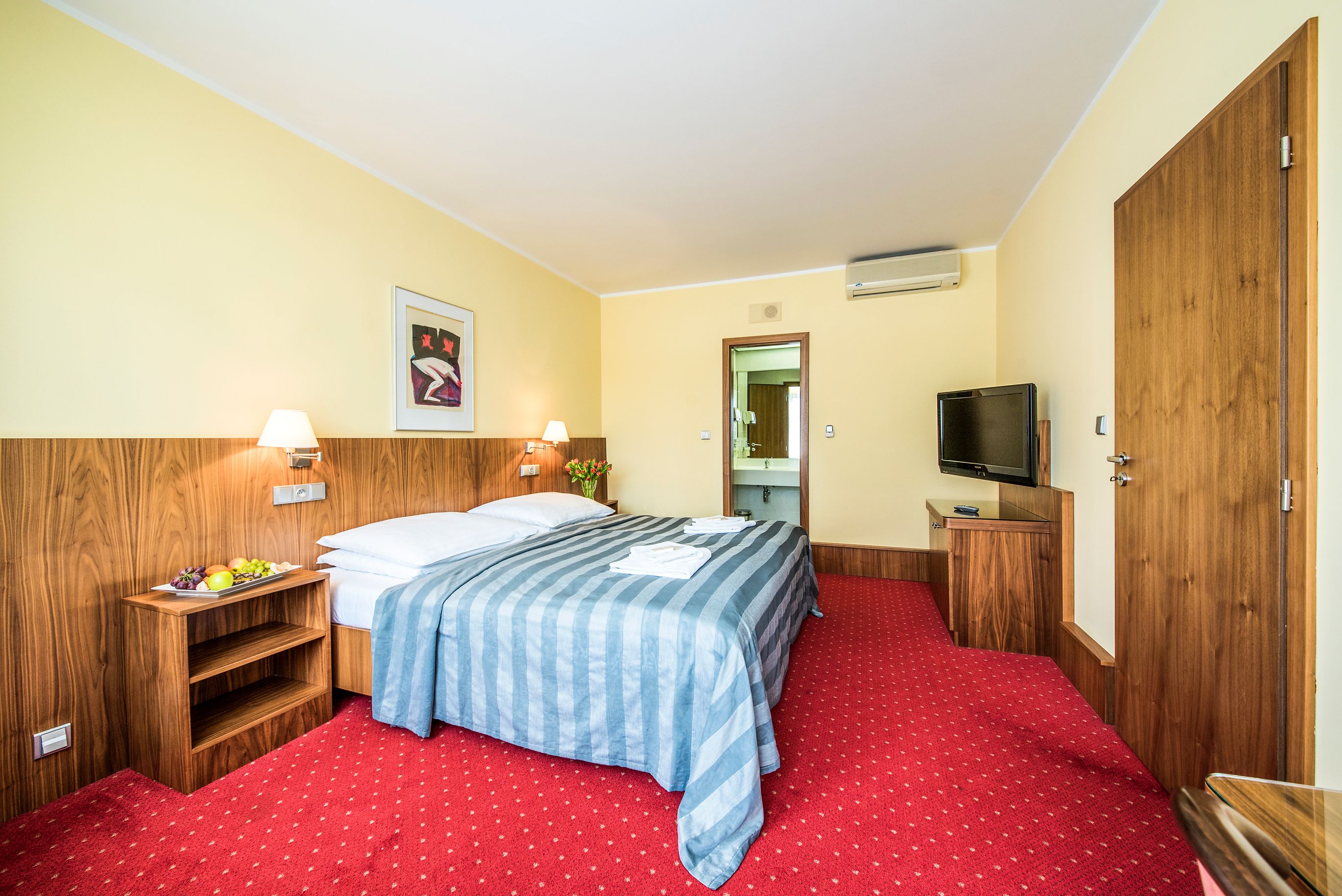 A hotel room with a bed, a TV, a wooden wardrobe, a bedside table with a lamp, and a small shelf with fruits.
