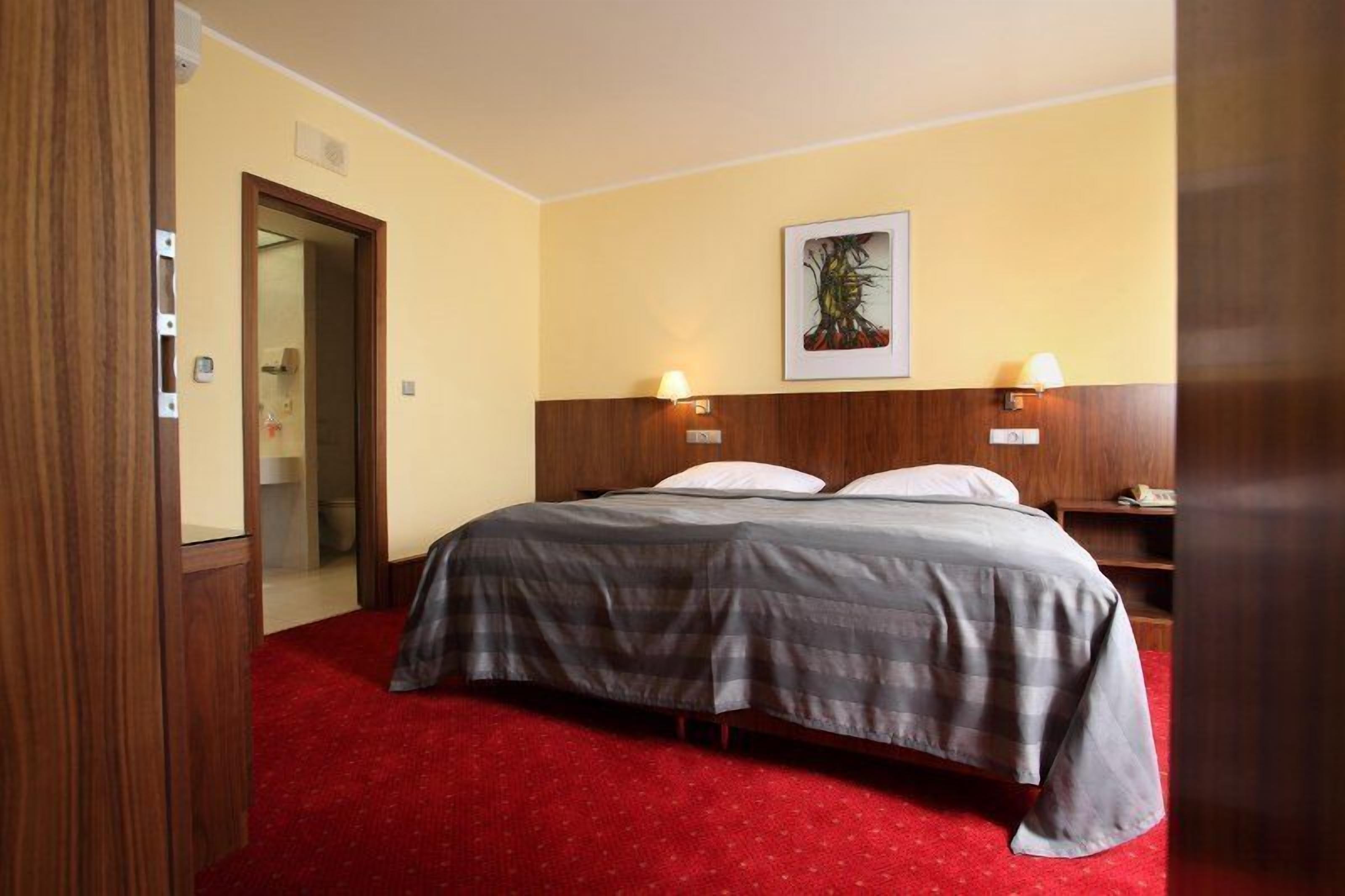 A hotel room with a bed, two bedside lamps, a framed artwork on the wall, and a red carpet.